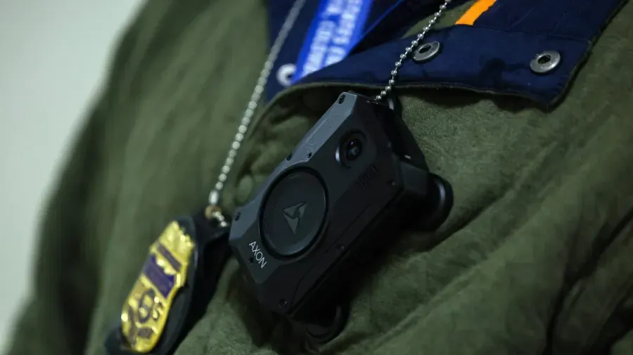 Close-up of an Axon body camera clipped to a green jacket, with a gold badge on a chain.