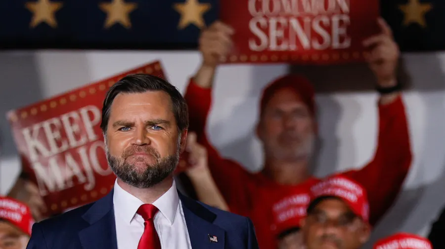Man in navy suit and red tie at rally, red 'Keep Majority' and 'Common Sense' signs behind him.