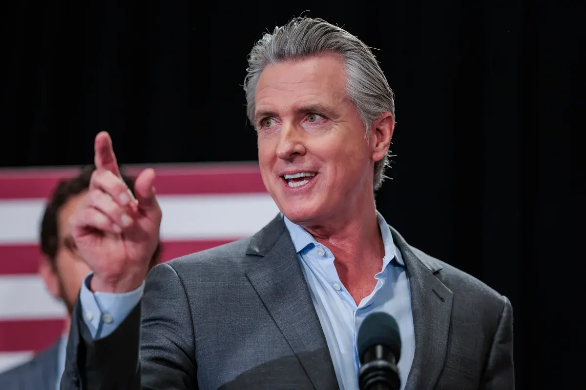 A man in a grey suit speaking at a microphone, pointing, in front of a striped flag backdrop.