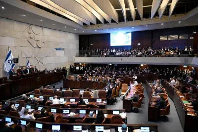 Wide view of Israeli parliament chamber with rows of seated members and Israeli flags.