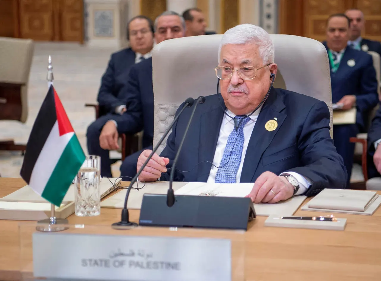 Older man in glasses and suit seated at a conference table beside a Palestinian flag and 'State of Palestine' placard.