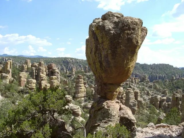 Tall balanced rock pinnacle amid eroded volcanic rock spires and green forest under blue sky.