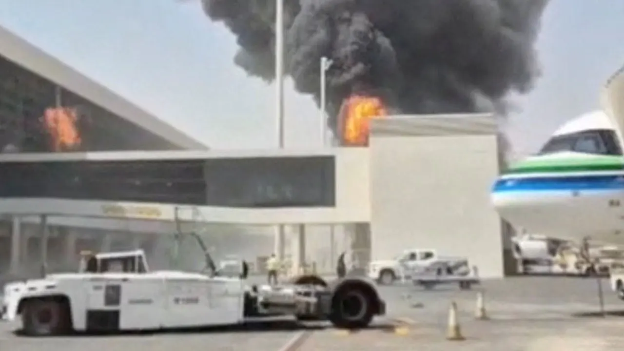 Smoke and fire rising behind an airport terminal beside a parked white passenger jet and tug.