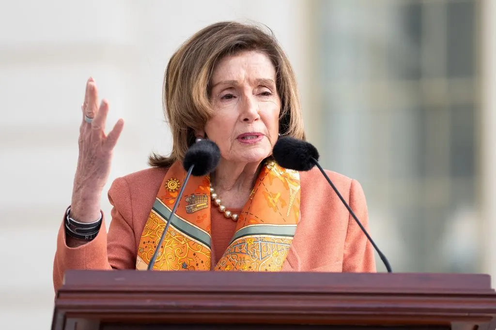 An older woman with shoulder-length hair speaks at a wooden podium with microphones, gesturing with one hand.