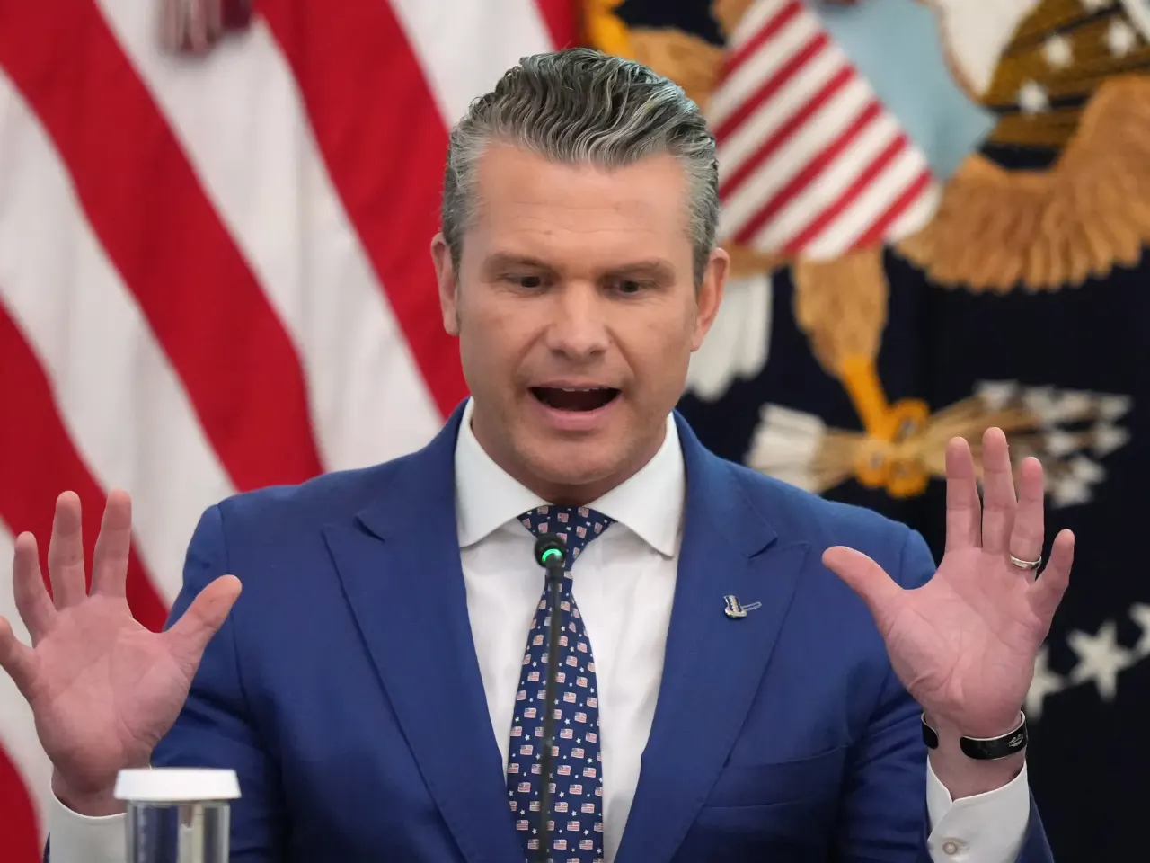 Man in blue suit speaking at a podium, hands raised, in front of US flag and seal.