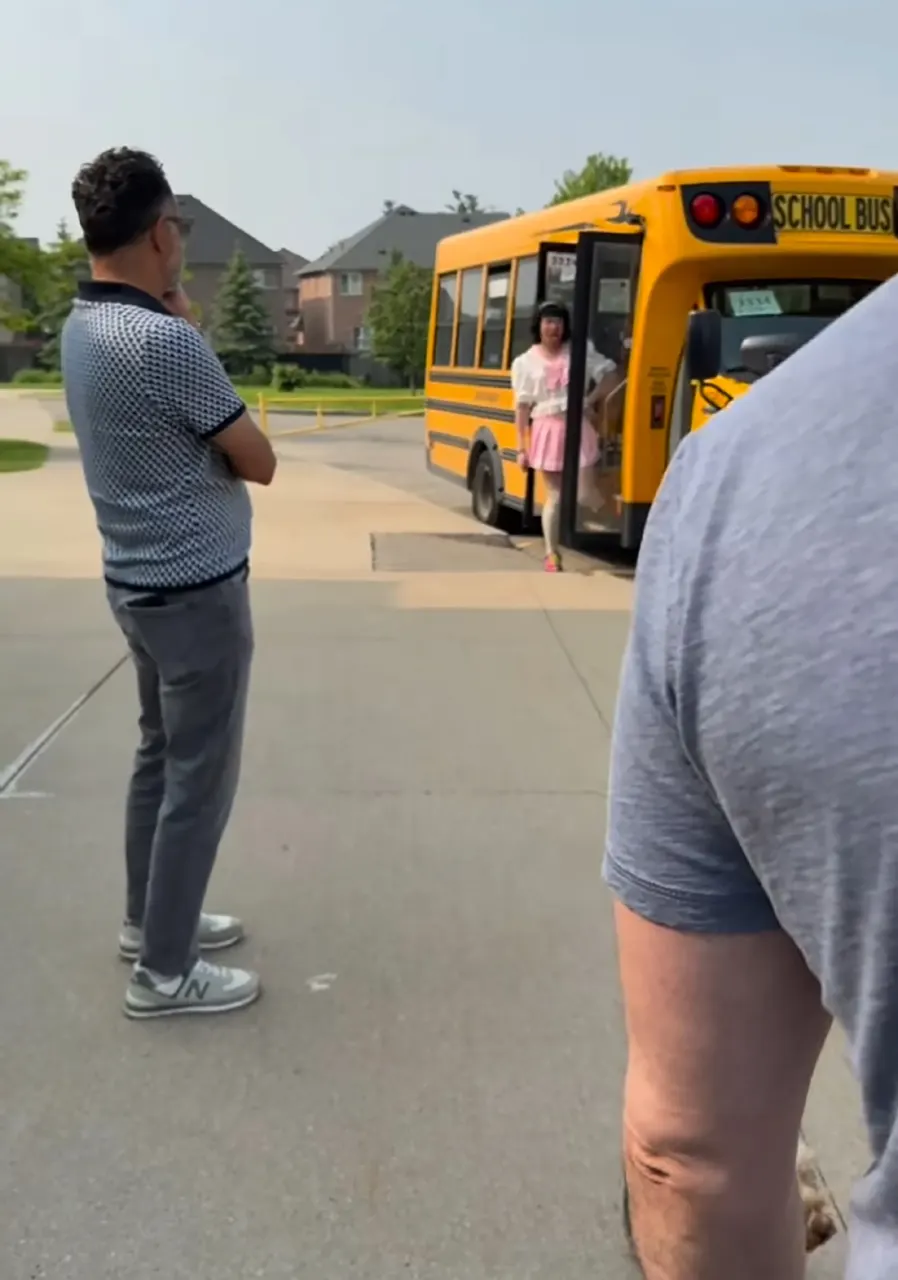 Yellow school bus with a child boarding at a curbside, with two adults watching nearby.