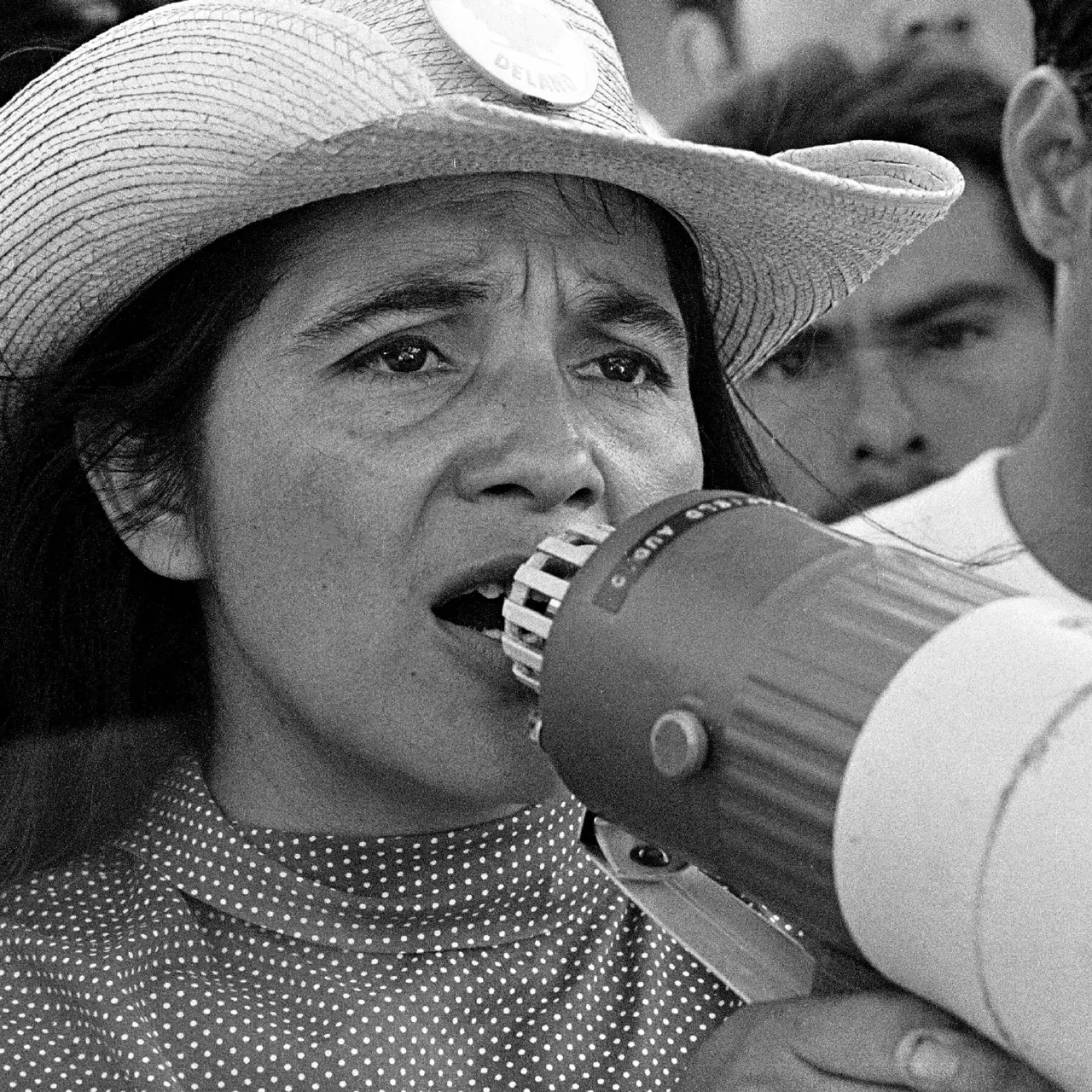 Black-and-white photo of a woman in a straw hat speaking into a megaphone at a rally.