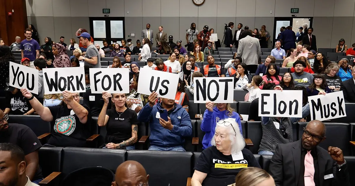 Crowd in council chamber holding handmade signs reading 'You Work For Us' and 'Not Elon Musk'.