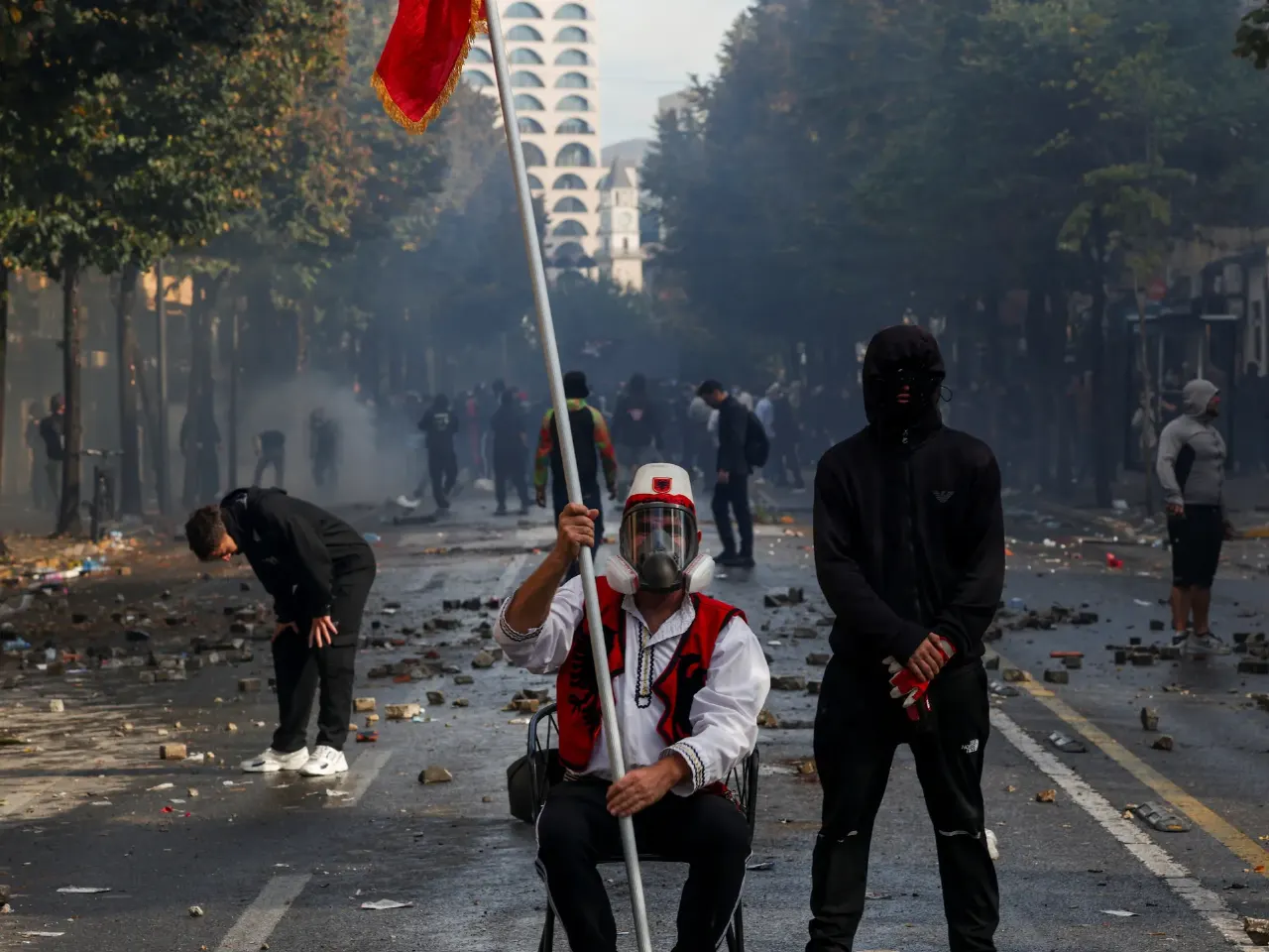 Protesters with red flag and masked figures amid smoke and debris on a city street